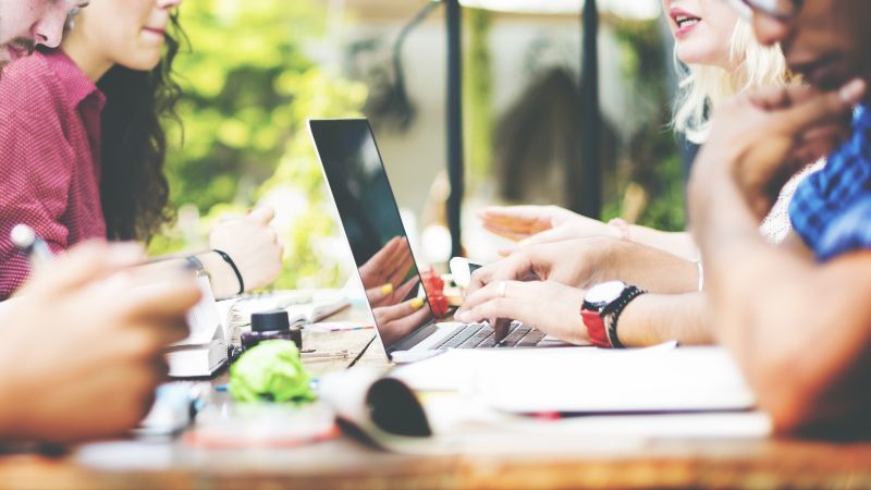 Zwei Personen mit einem Laptop sitzen weiteren Personen gegenüber 