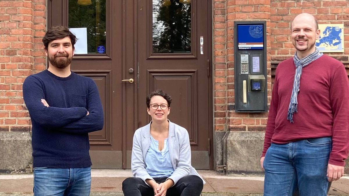 Kristopher Söderström (PhD) und Nicolas Rüffin stehen vor der Eingangstür eines alten Universitätsgebäudes. Auf den Treppenstufen in der Mitte sitzt Dr. Katharina C. Cramer
